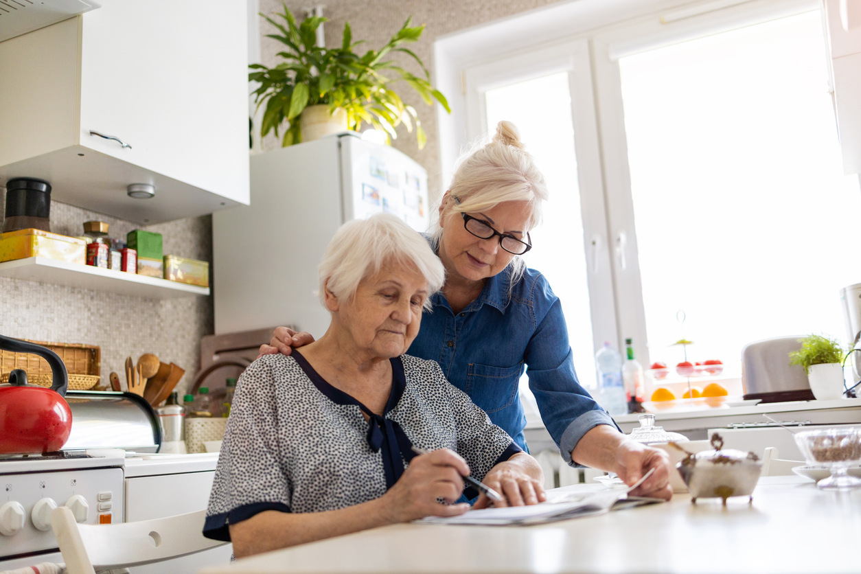 An elderly woman sitting at a table looking over estate planning paperwork while her adult daughter stands at her side.